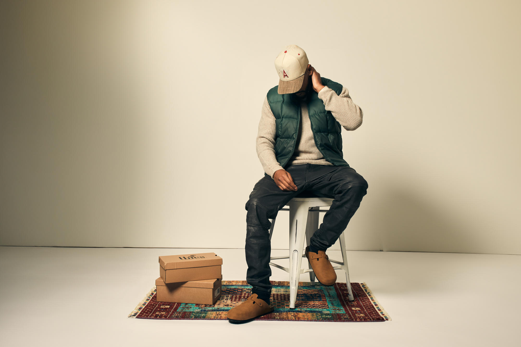 A model seated beside Locals Only shoe boxes on a warm-toned rug in studio — evoking the moment of receiving and authenticating a Locals Only collectible.